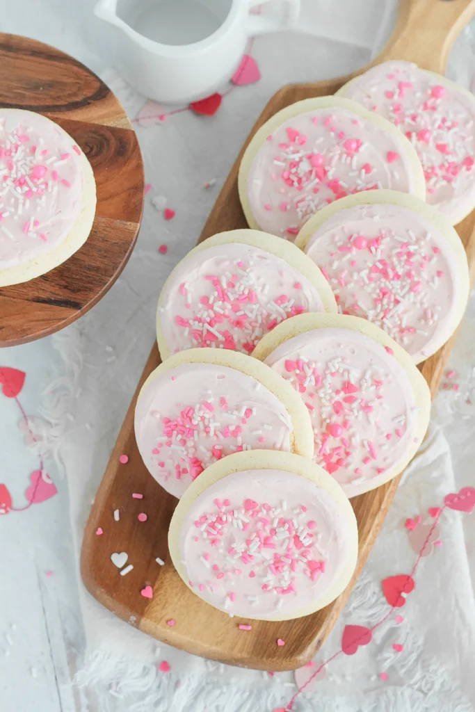 Sept biscuits déposés sur une planche de bois avec des coeurs dans l'arrière plan. Les biscuits sont couvert d'un glaçage rose.