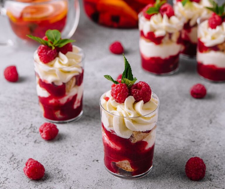 Bagatelle dans un contenant en verre sur une surface grise couvert de quelque framboise. Le contenant contient du gâteau à la vanille, un coulis de framboise et une glaçage blanc